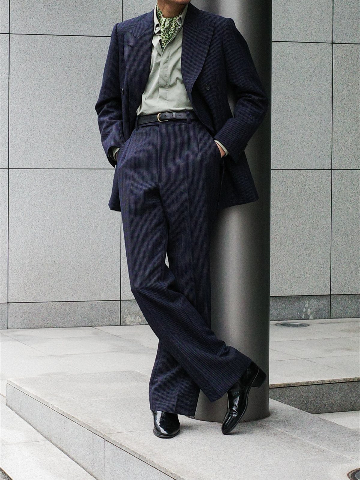 Man in a navy pinstripe suit and green shirt leaning against a grey pillar.