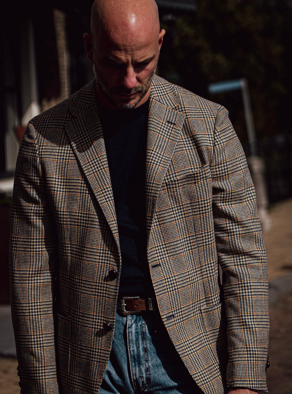 A man wearing a brown plaid blazer, dark shirt, and blue denim jeans outdoors.