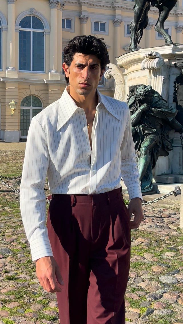 Man in a white striped shirt with large collars and burgundy trousers standing outdoors.