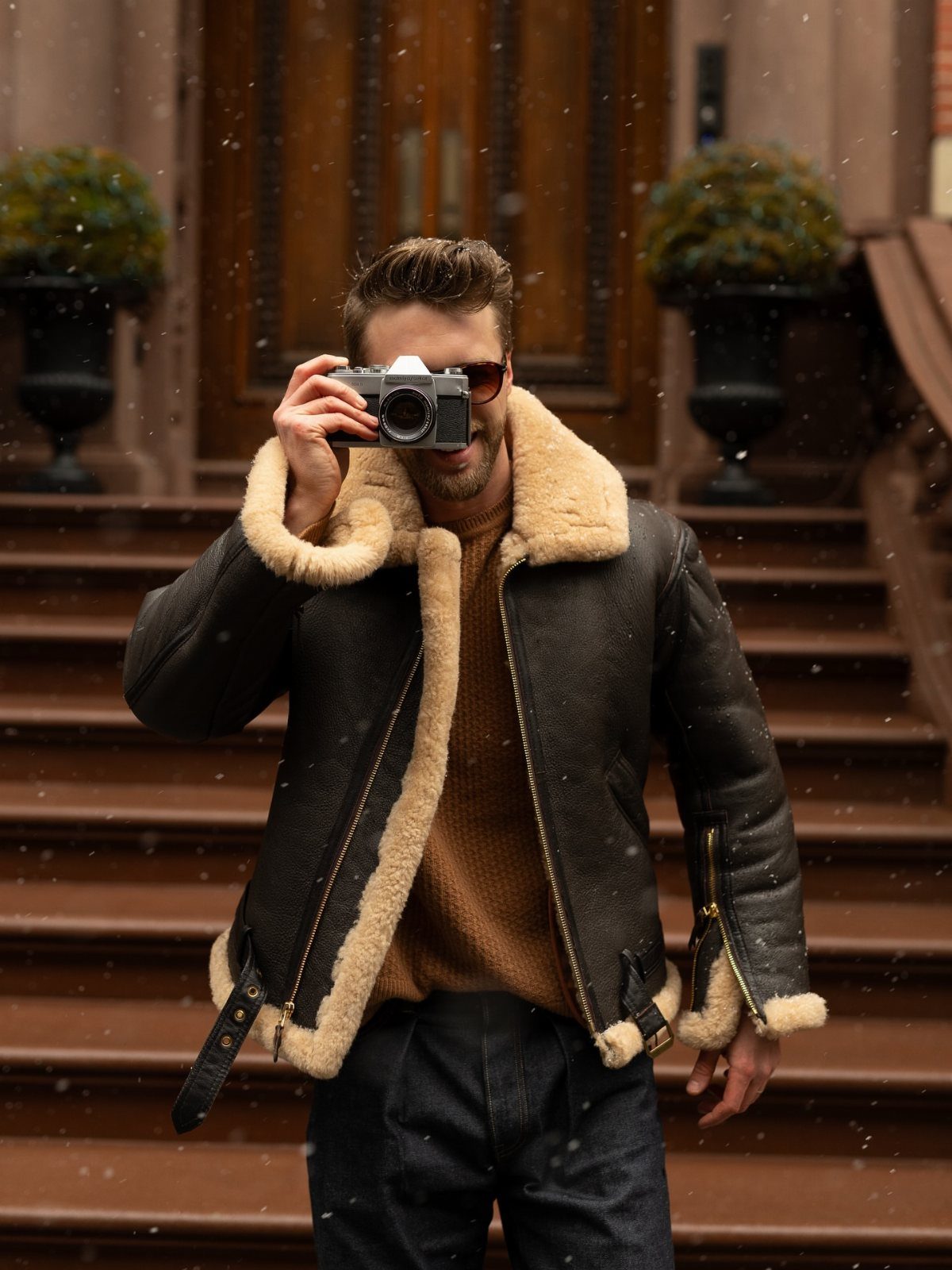 Man in a dark leather shearling jacket holding a vintage camera on outdoor stairs.