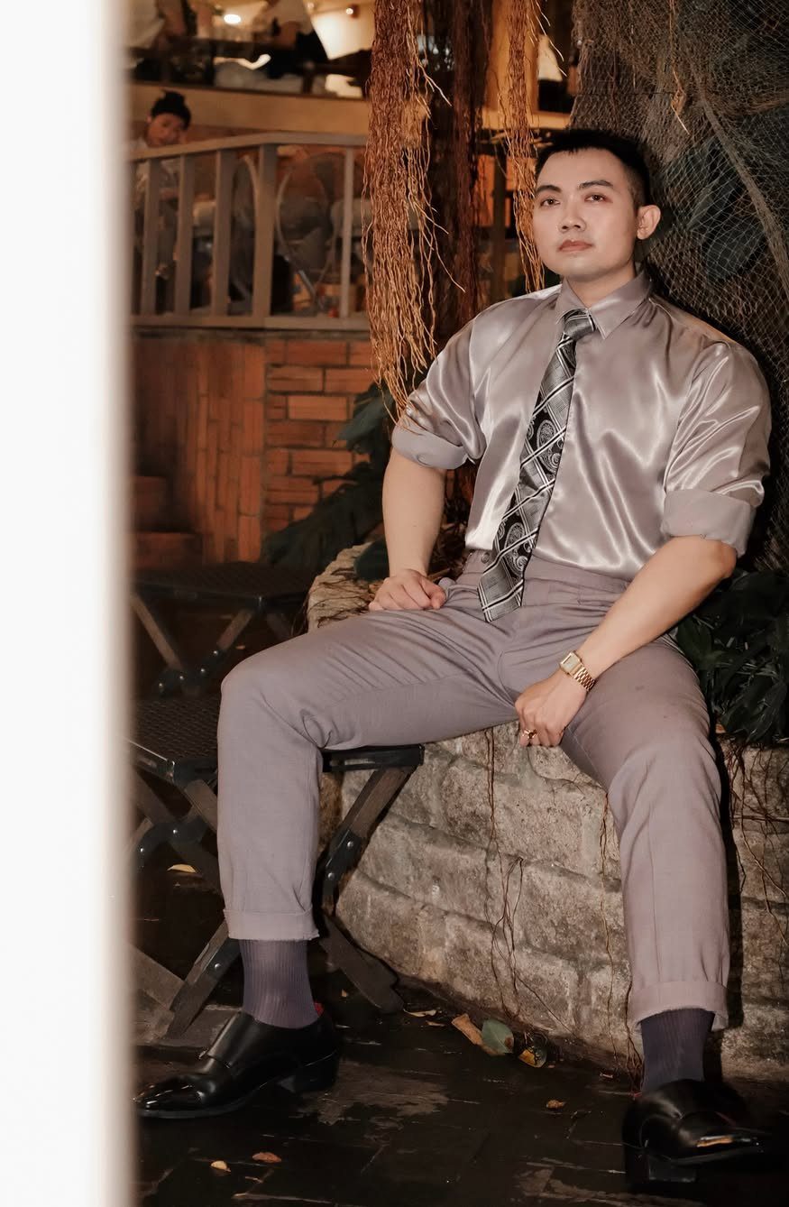 A man wearing a shiny grey satin shirt and patterned tie sitting on a stone ledge.