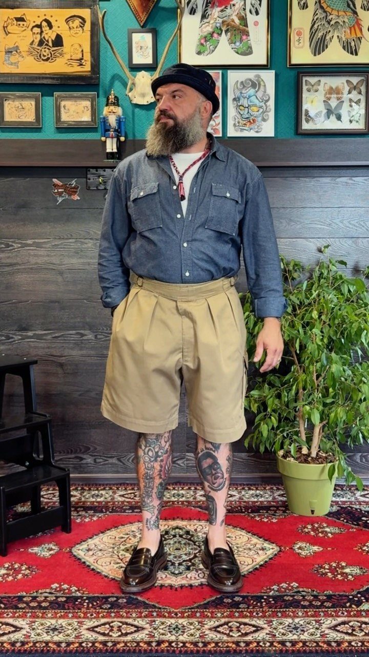 Bearded man in a blue button-down shirt and tan pleated shorts standing on a red rug.
