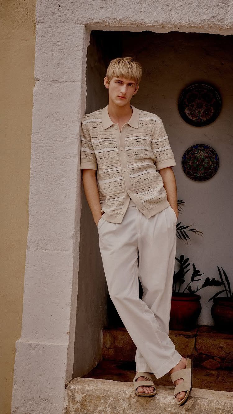 A young man wearing a beige textured shirt and white casual trousers leaning against a stone doorway.