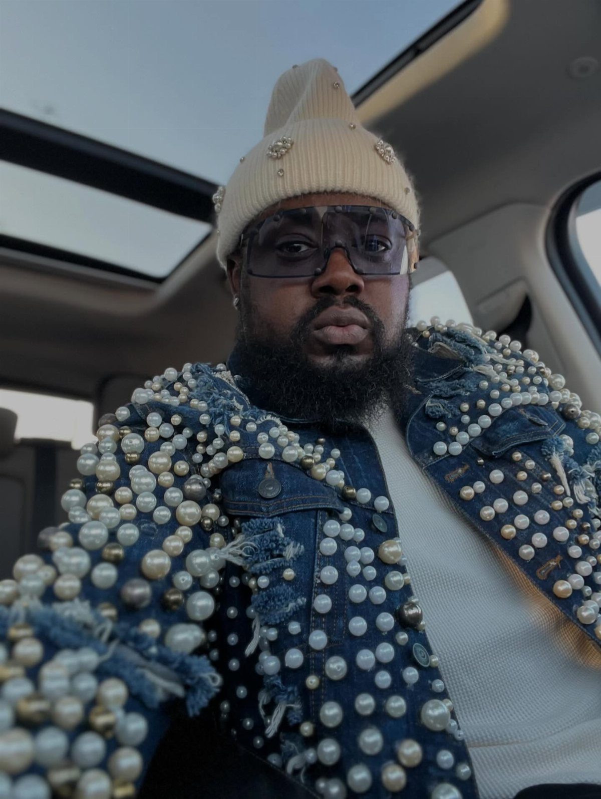 Man with a beard wearing a pearl-embellished denim jacket and a cream beanie inside a car.