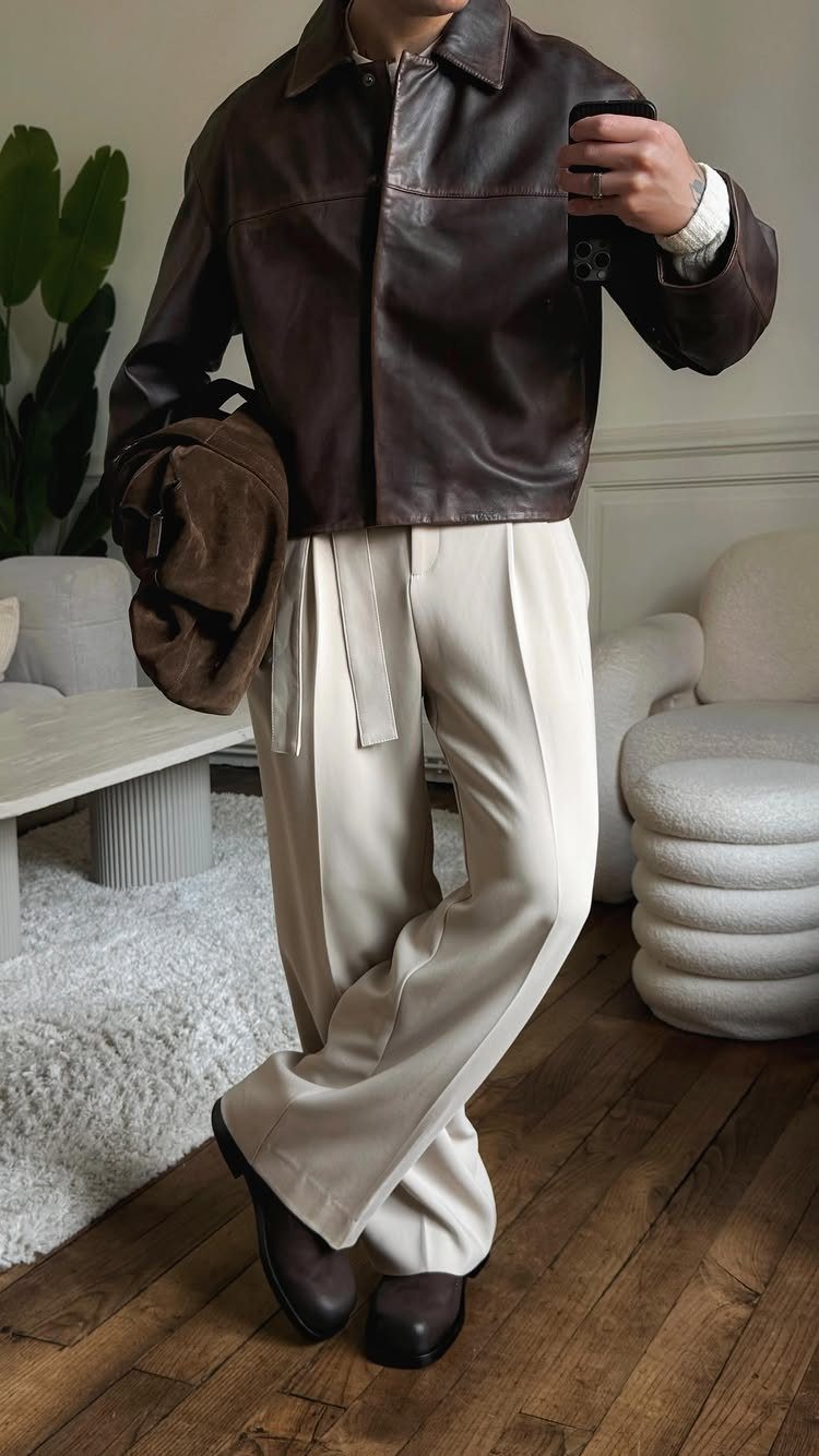 Man wearing a dark brown leather jacket and cream wide-leg trousers standing in a room.