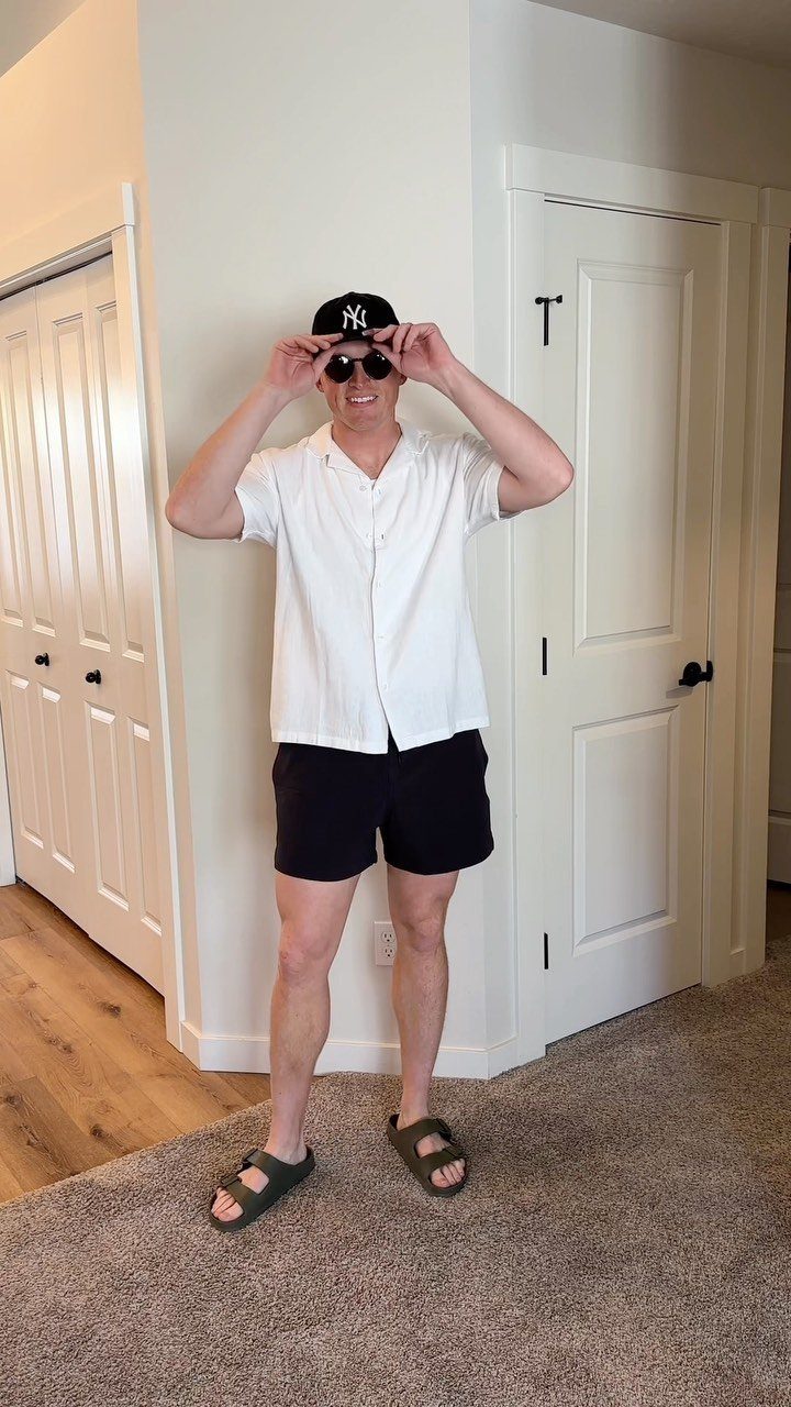 Man wearing a white linen camp-collar shirt, black shorts, and a baseball cap indoors.