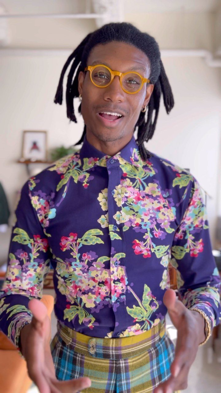 Man with dreadlocks and yellow glasses wearing a purple floral patterned shirt and plaid trousers.