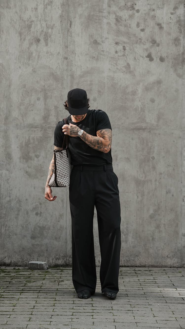 Man in all black outfit with wide-leg trousers and a patterned bag standing against a concrete wall.
