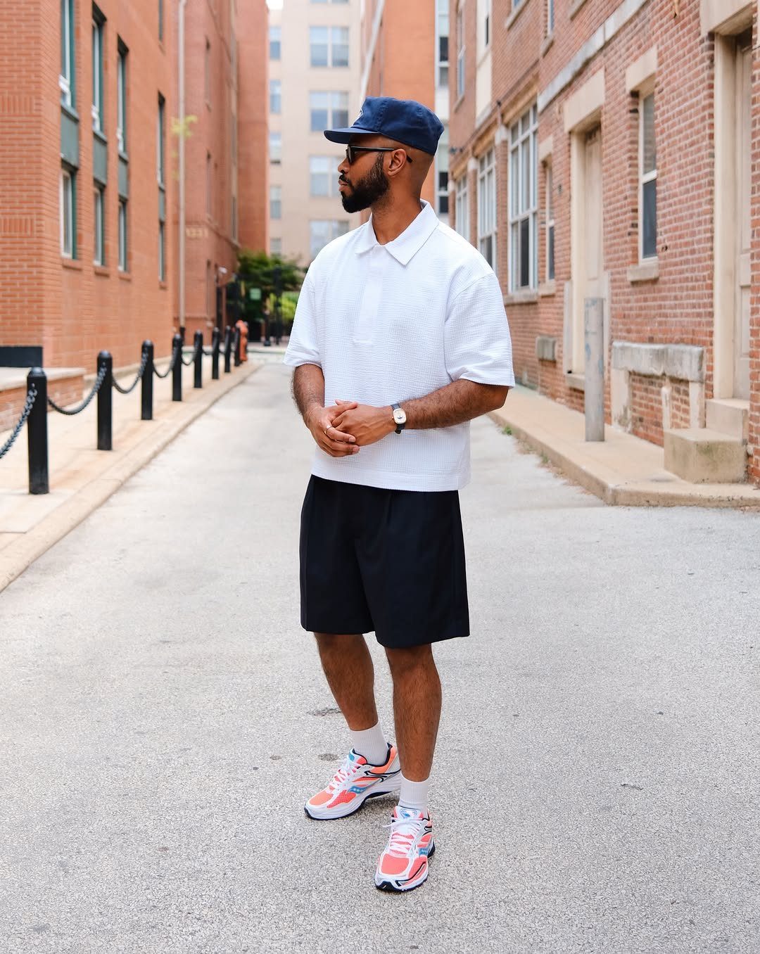 Man in white polo, black shorts, and colorful sneakers standing in a brick alleyway.