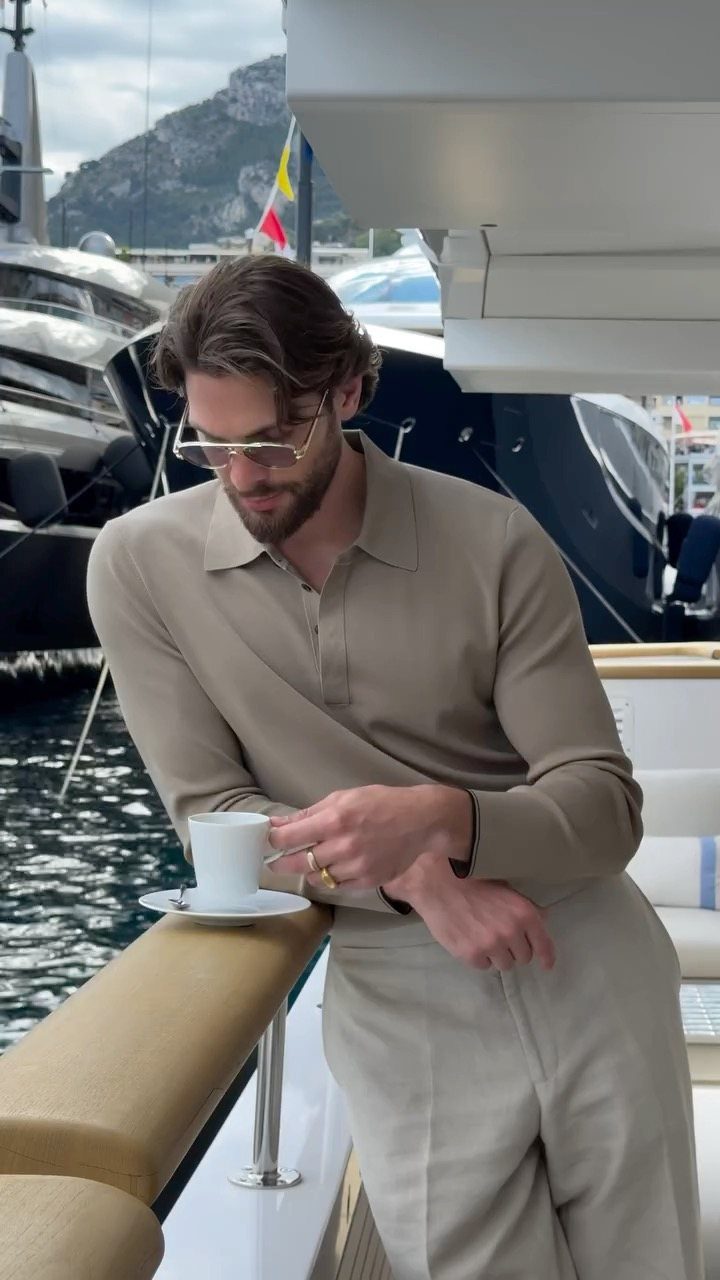 Man in beige long-sleeved polo and trousers leaning on a yacht railing with coffee.