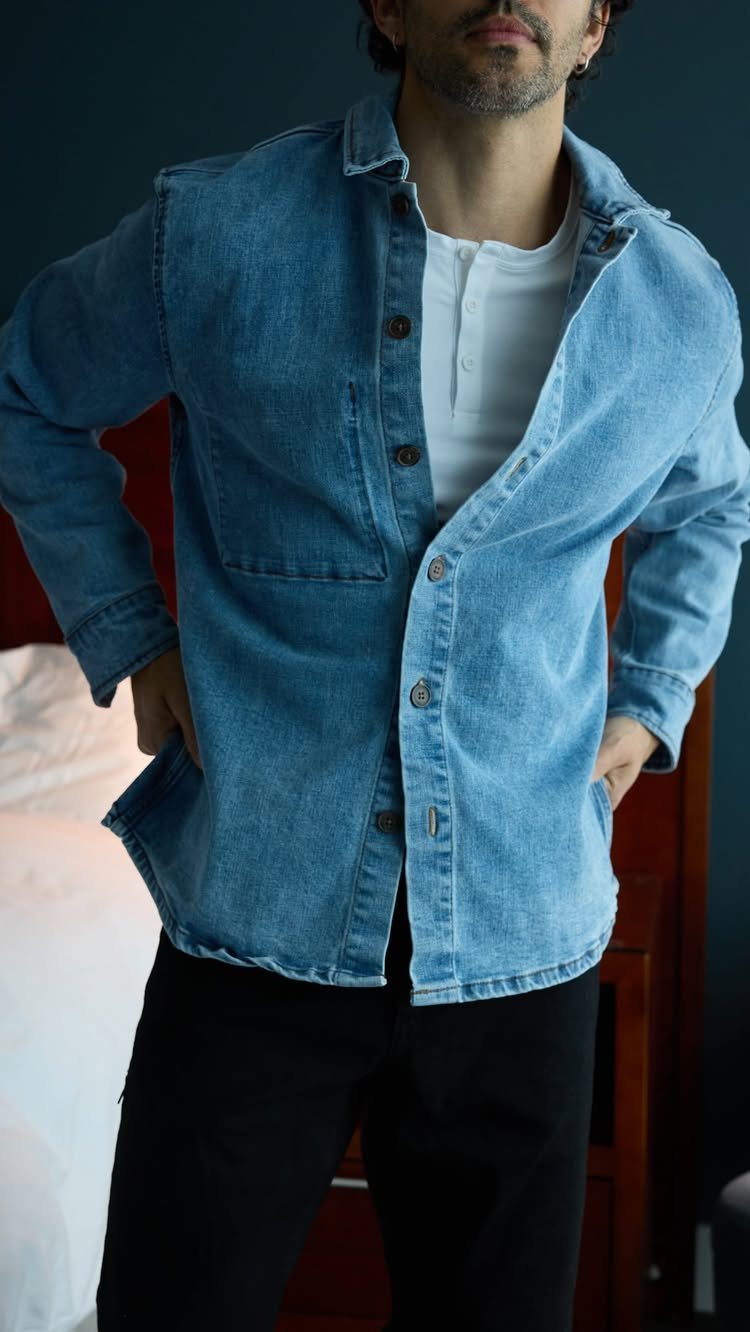 Man wearing a light blue denim shirt over a white henley and black pants.