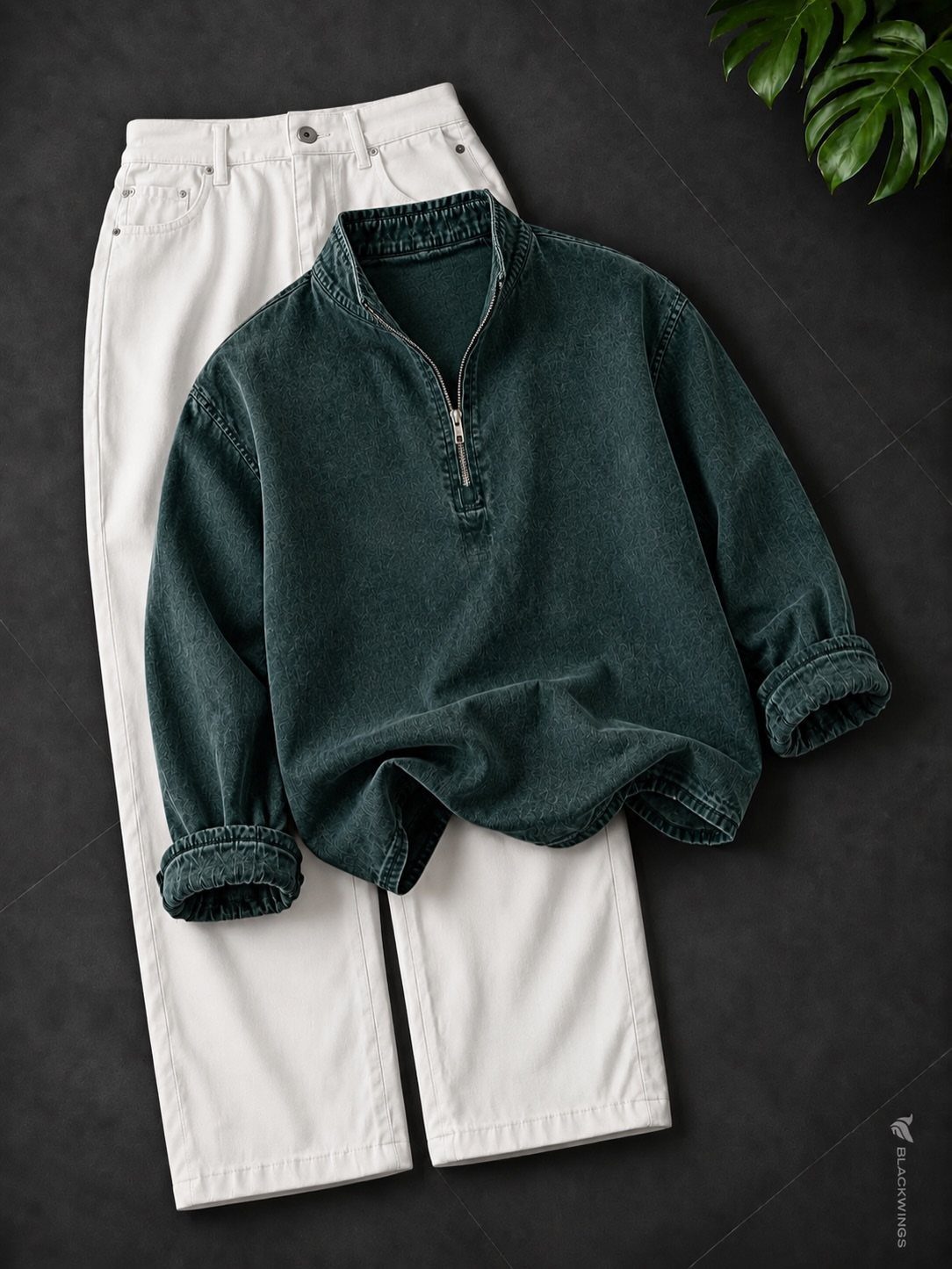 A dark green quarter-zip pullover shirt laid on top of white trousers on a dark background.