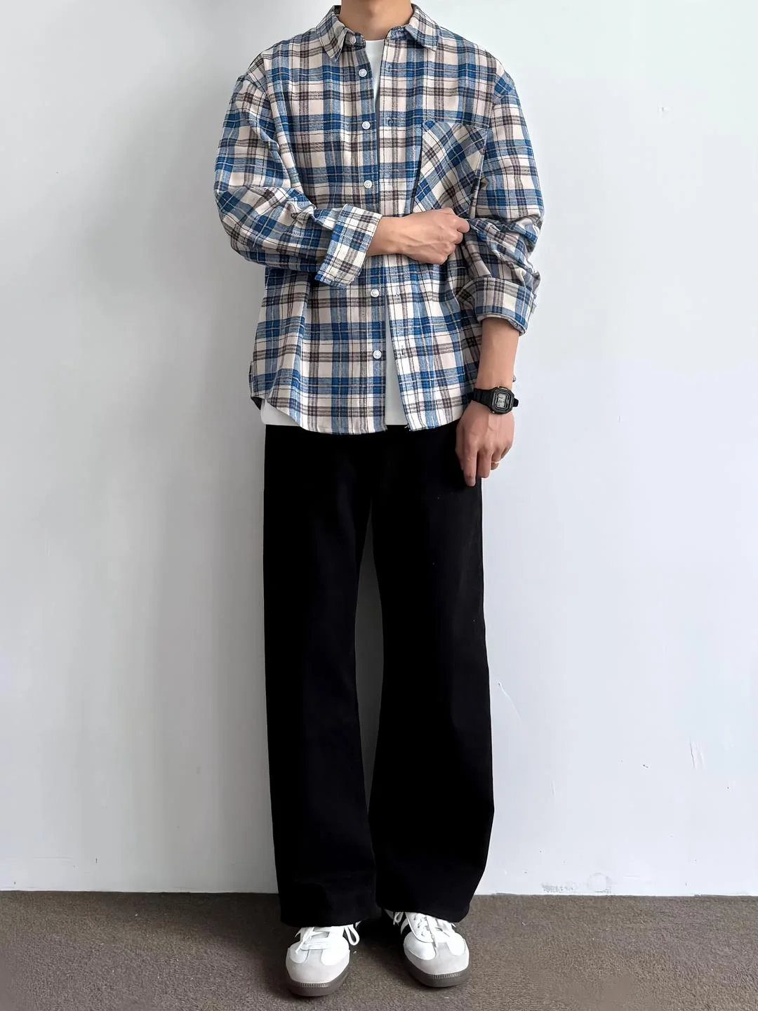 Man wearing a plaid shirt, wide black trousers, and white sneakers against a white wall.