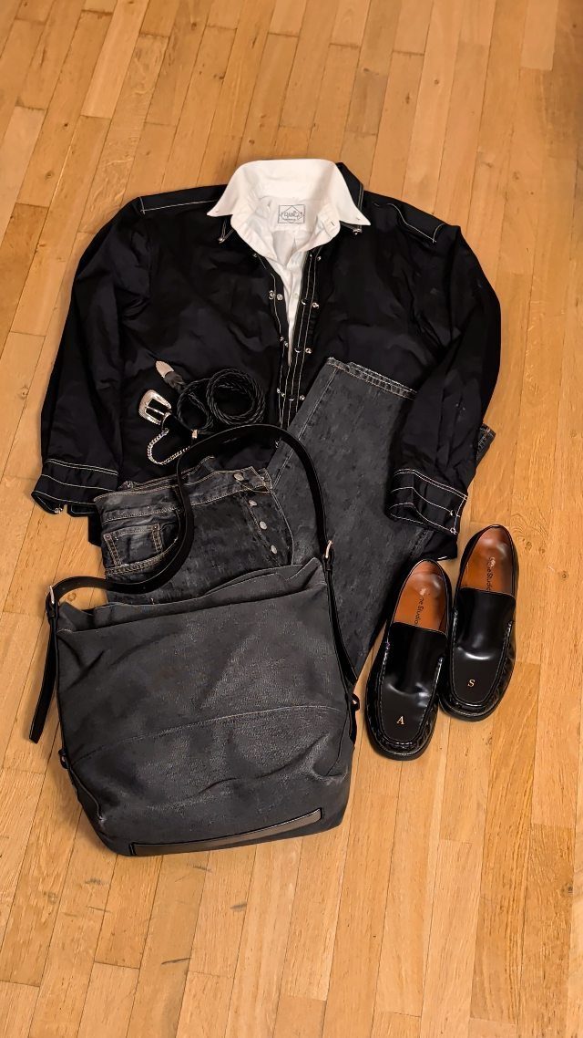 Flat lay of a black jacket, white shirt, denim jeans, grey bag, and black loafers on wood.