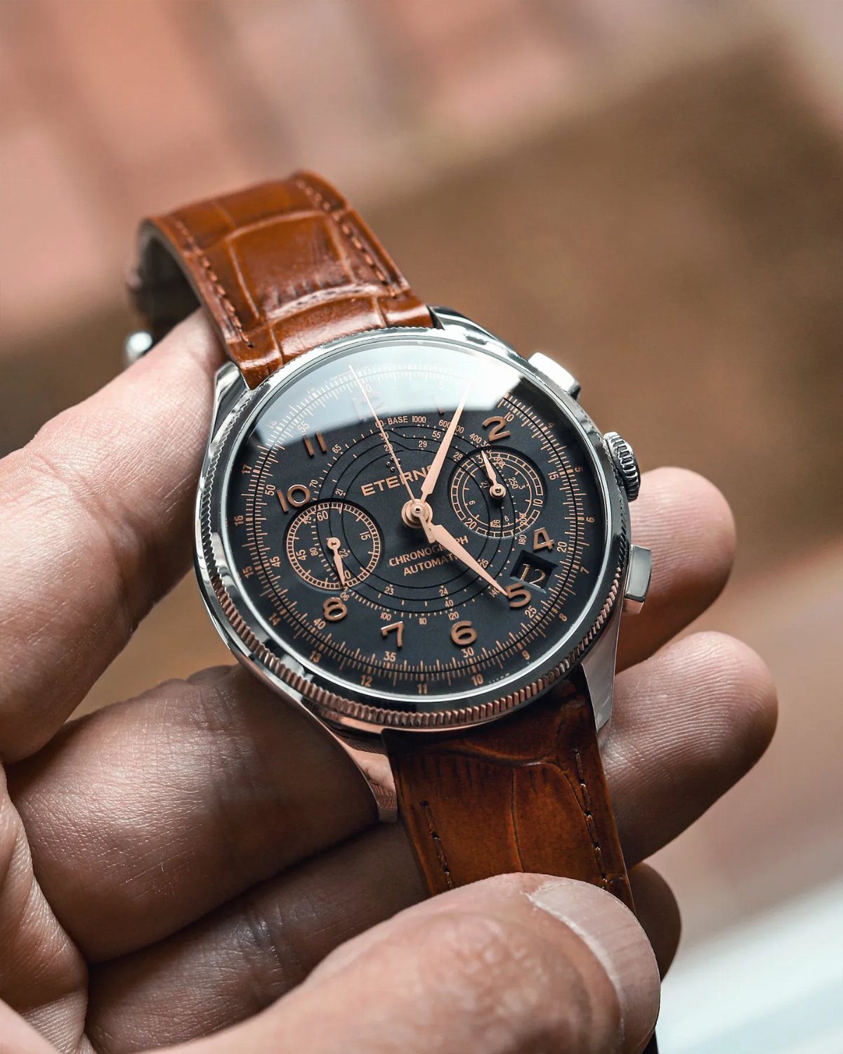 A silver chronograph wristwatch with a black dial and brown leather strap held in a hand.