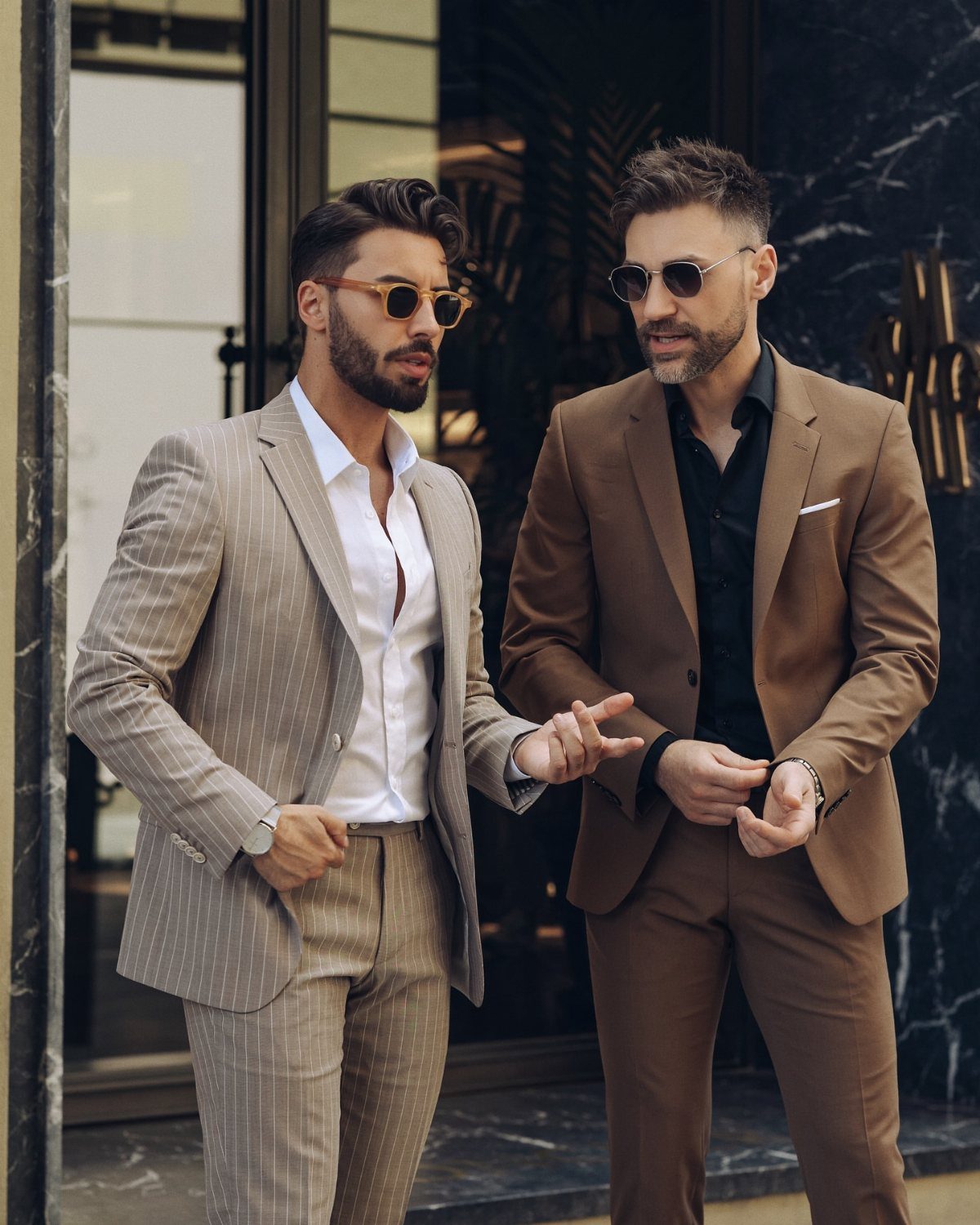 Two stylish men wearing tailored suits and sunglasses standing in front of a marble building.