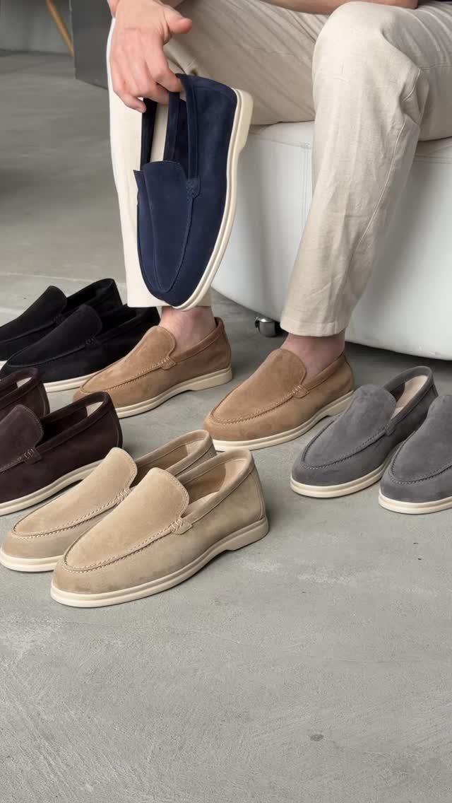 A person holding a navy loafer surrounded by various colored suede loafers on a floor.