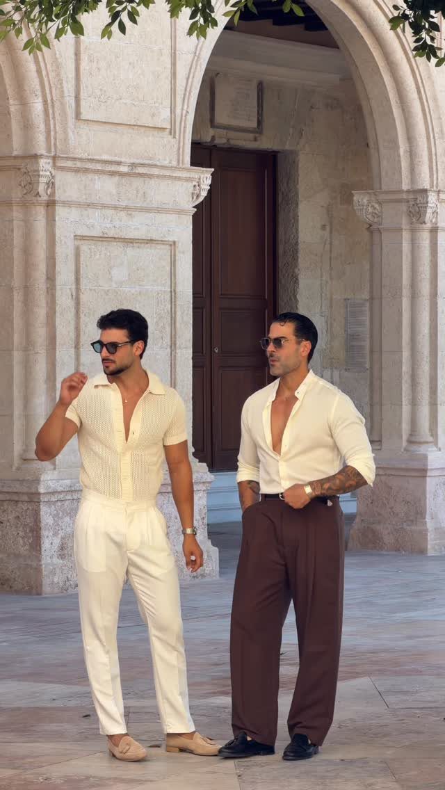 Two men posing in elegant summer fashion standing before classical stone arches.