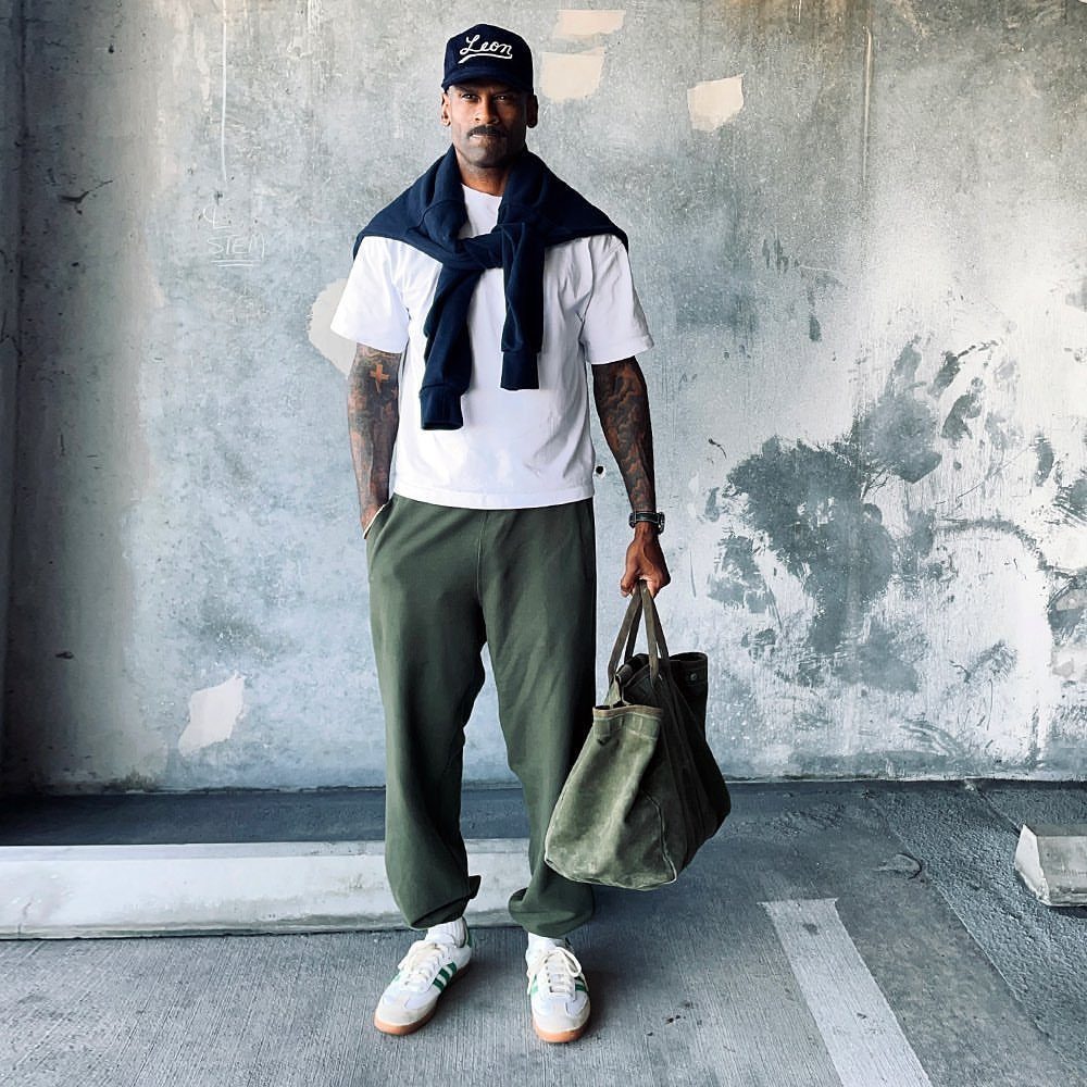 Man in white t-shirt, olive joggers, and navy sweater standing against a concrete wall.