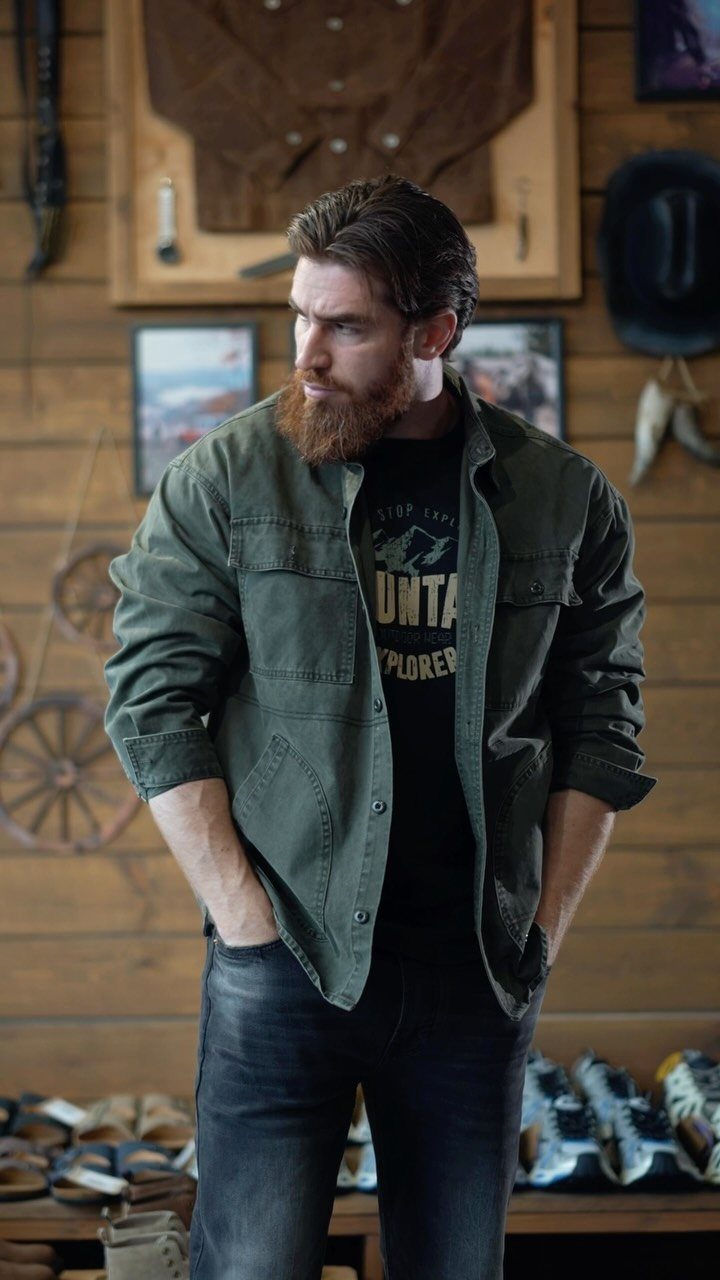A bearded man wearing an olive green shirt and black graphic t-shirt standing indoors.
