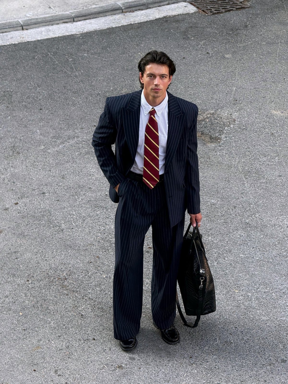A young man posing in a navy pinstripe suit and striped tie while holding a black bag.