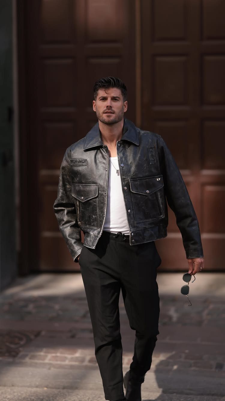 A man walking in a black leather jacket, white tank top, and black trousers.