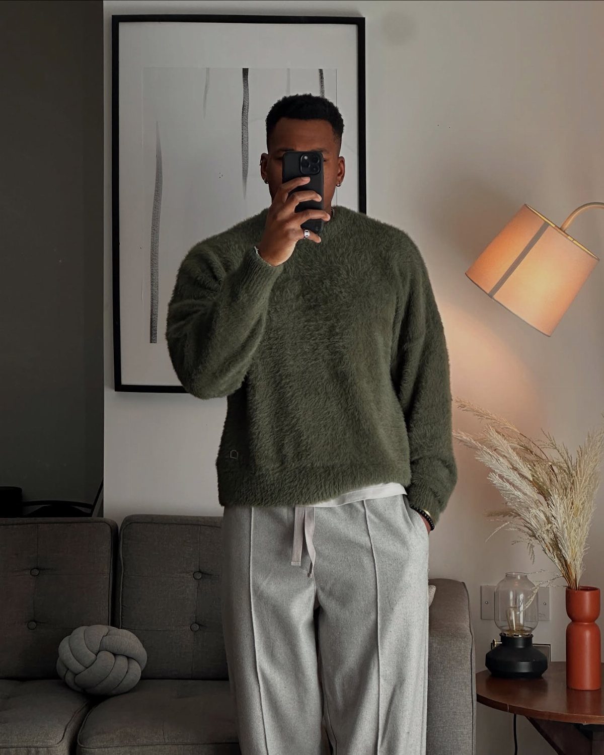Man in olive green fuzzy sweater and grey trousers taking a mirror selfie indoors.