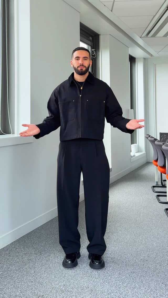 Man standing in an office hallway wearing a black cropped jacket and wide-leg trousers.