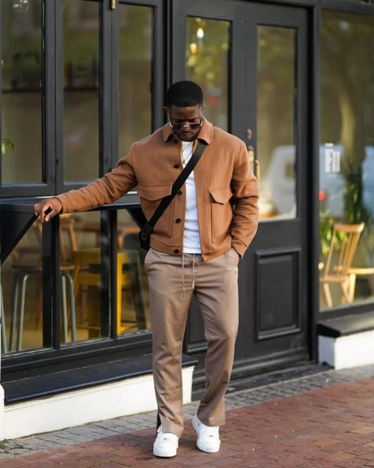 Man wearing a brown jacket, white t-shirt, tan trousers, and white sneakers standing outdoors.
