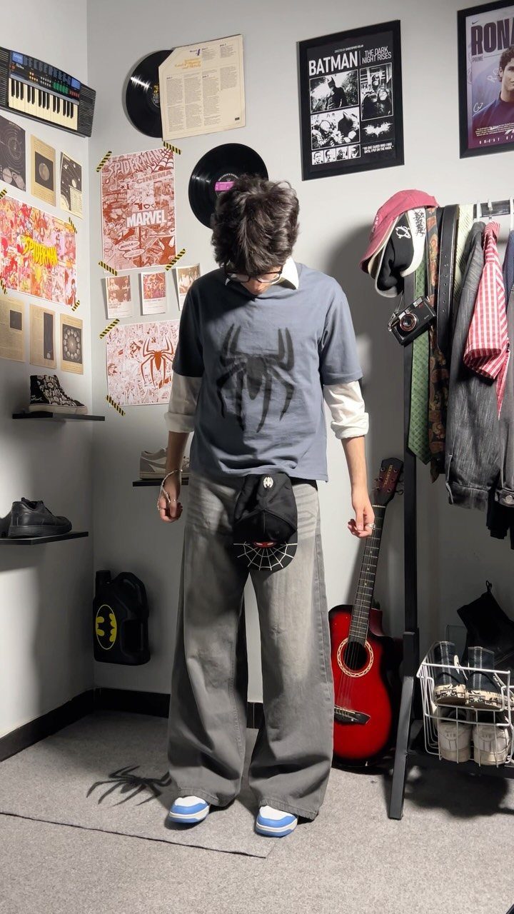 A young man wearing a graphic t-shirt and wide-leg grey trousers stands in a room.