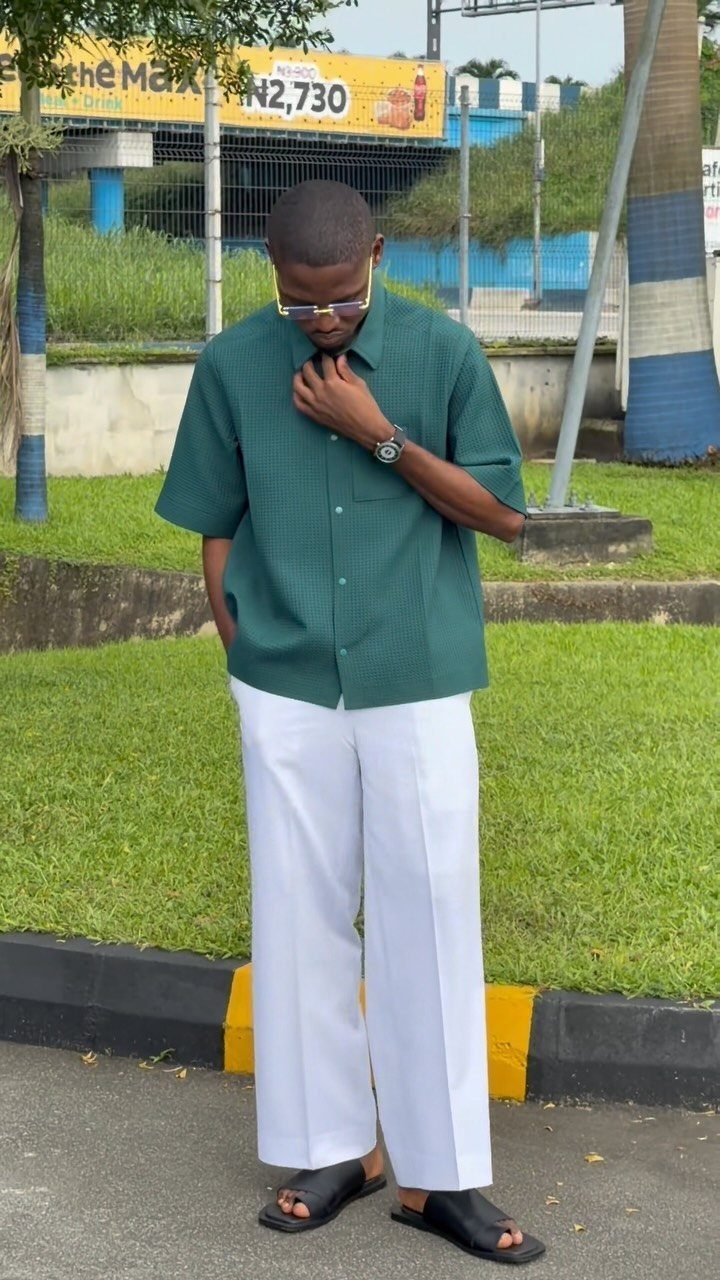 A man wearing a green short-sleeved shirt and wide white trousers standing outdoors.