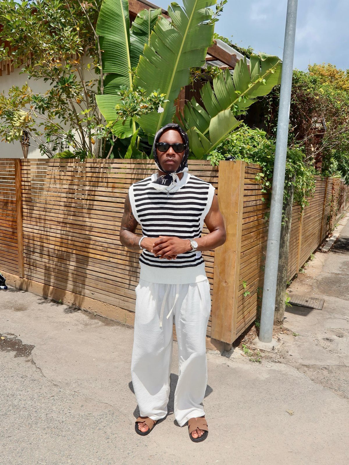 Man wearing striped sleeveless top, white linen pants, and sunglasses standing by a fence.