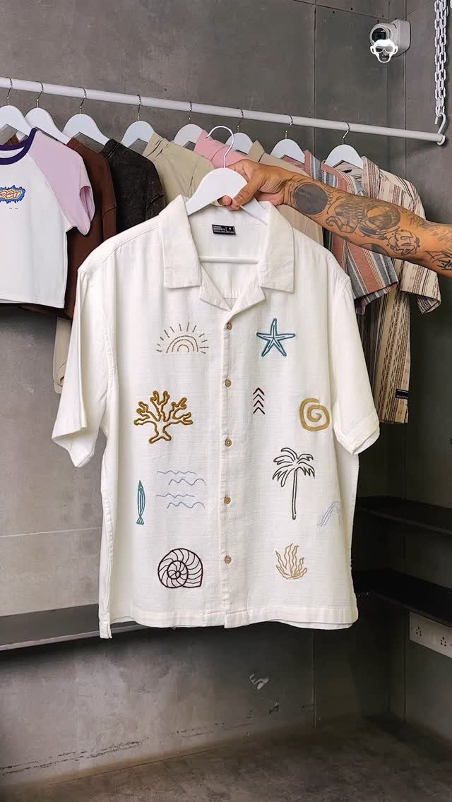 A person holding a white shirt with embroidered beach motifs against a clothing rack.