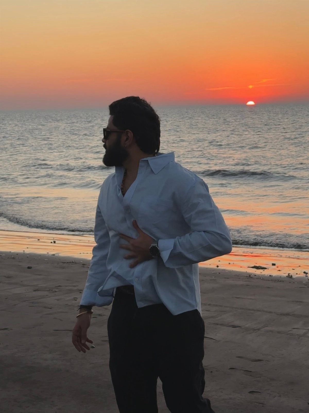 A bearded man in a light blue shirt stands on a beach during a vibrant sunset.