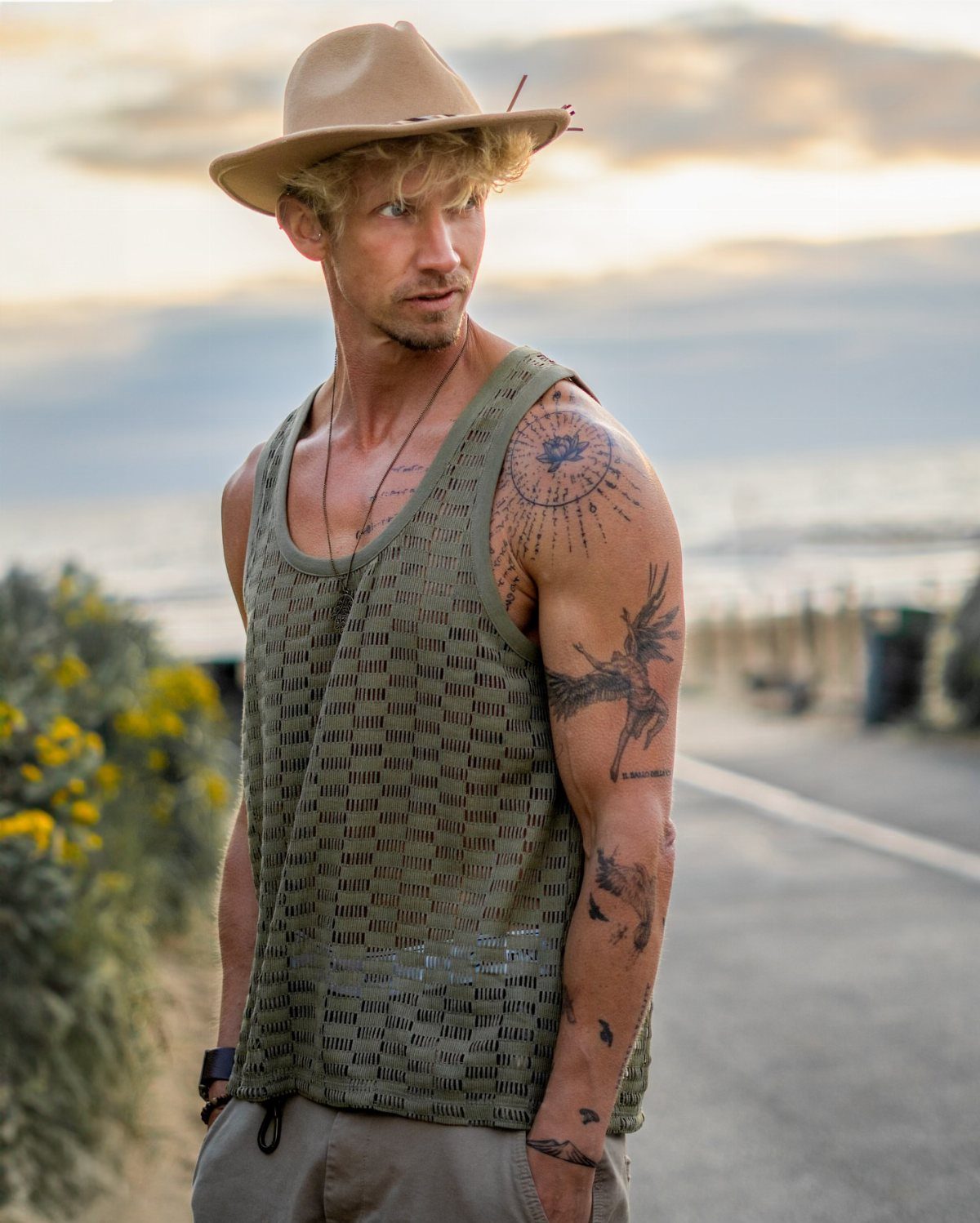 A man in a mesh tank top and fedora standing by a coastal road.