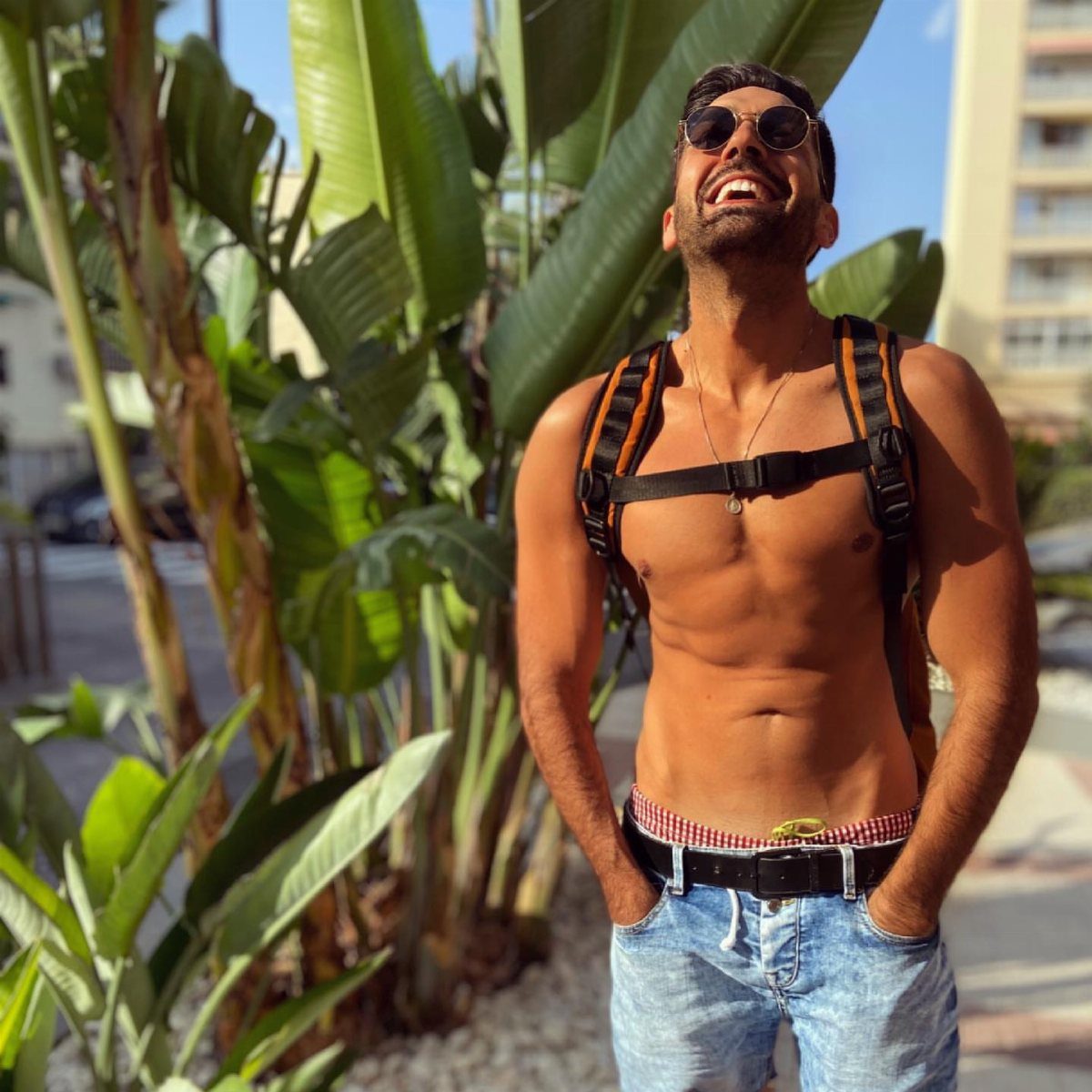 A shirtless man wearing sunglasses and a backpack smiles while standing near tropical plants.