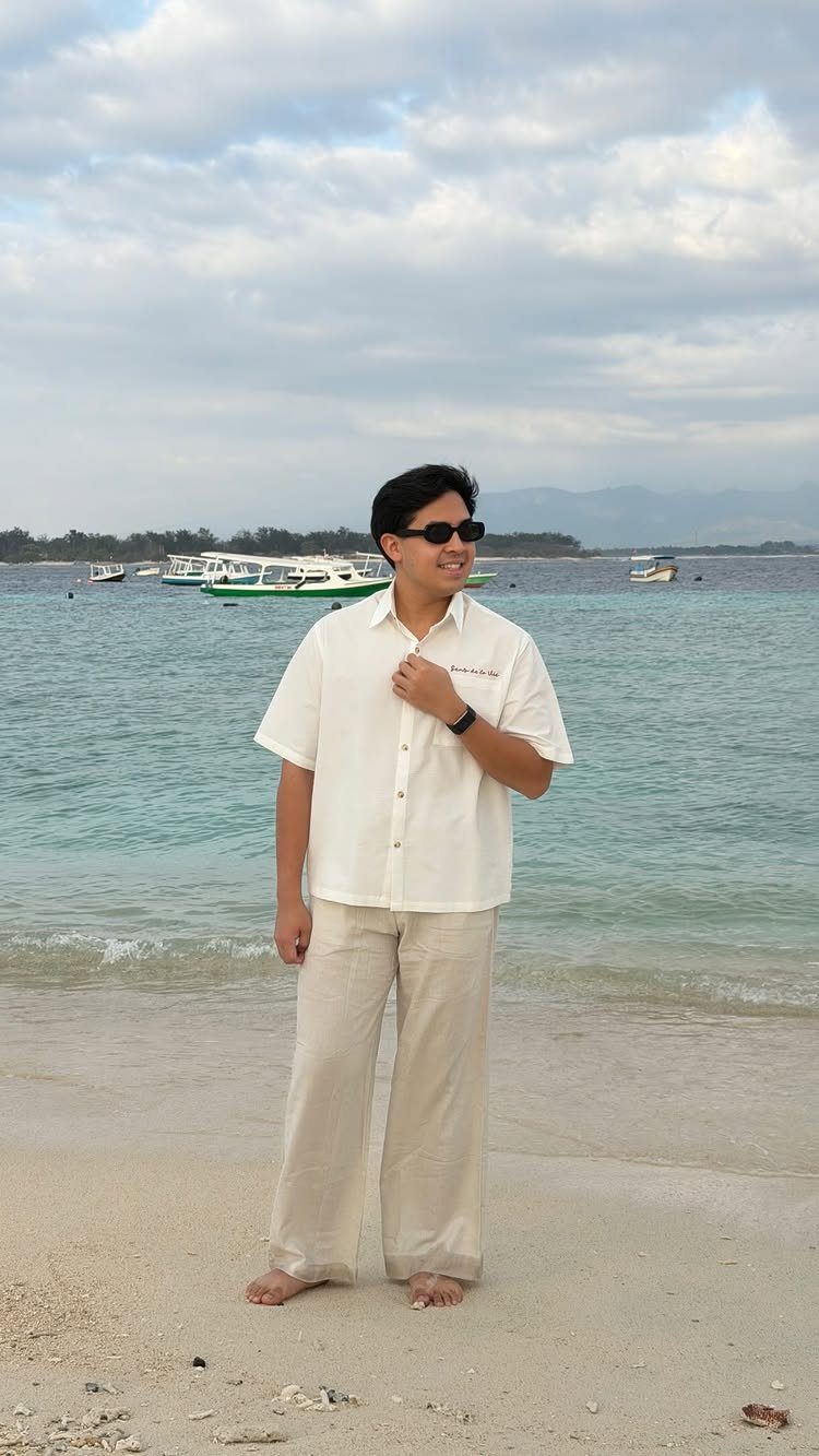 Man in white linen shirt and beige trousers standing on a tropical beach.