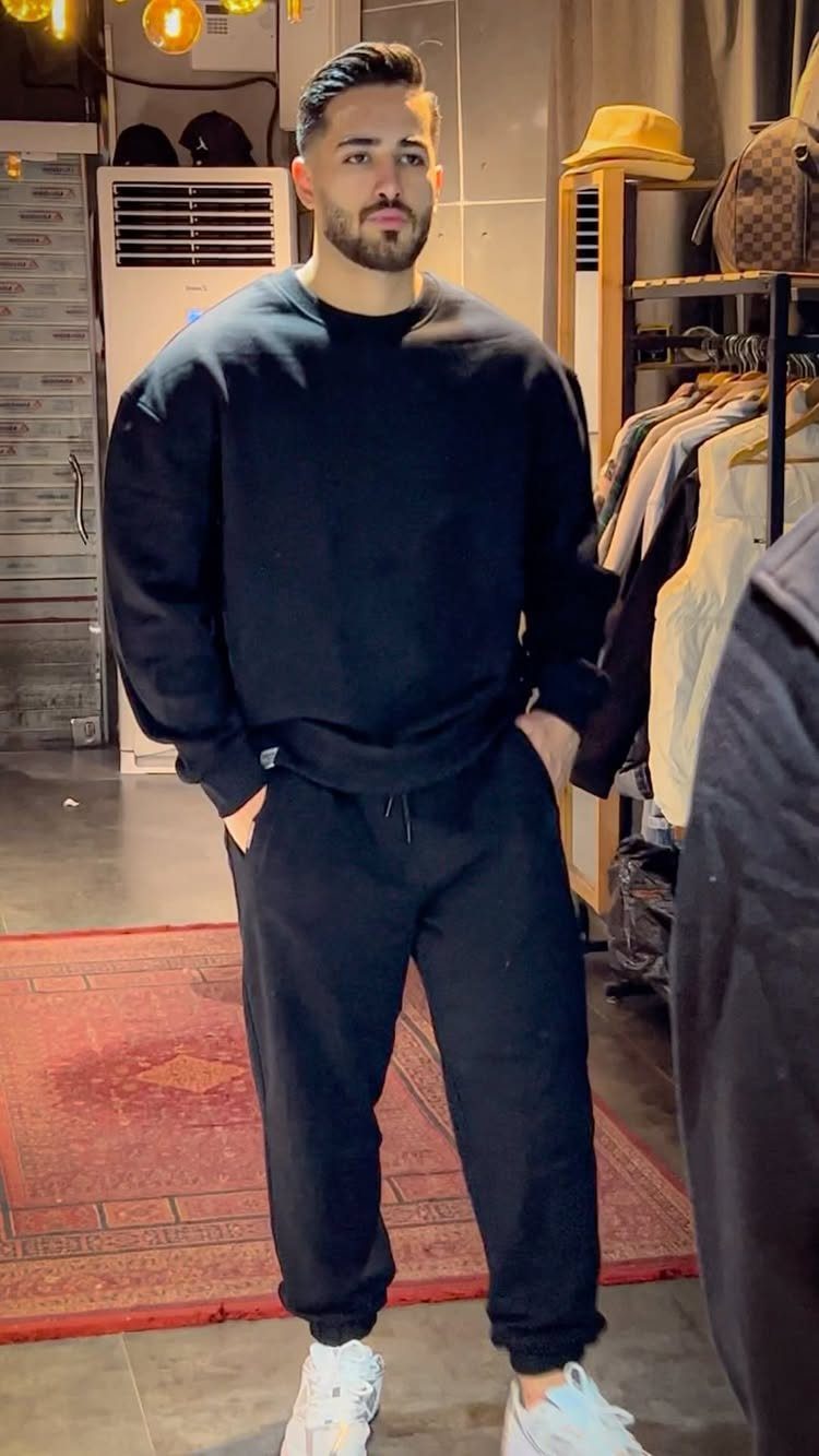 A man wearing a black sweatshirt and matching sweatpants with white sneakers standing in a shop.