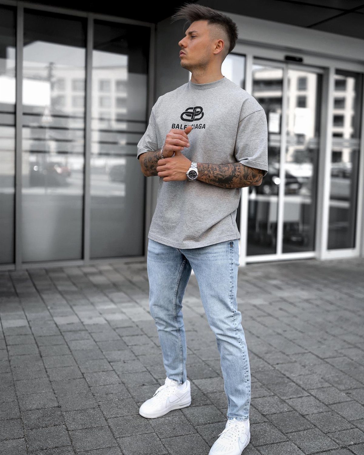 Man in a grey t-shirt and light blue jeans standing on a paved walkway.