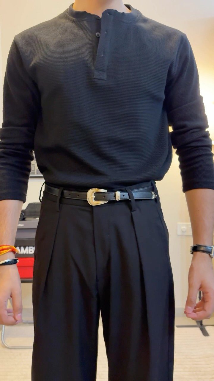 Man wearing a black textured henley shirt and black pleated trousers with a gold buckle belt.
