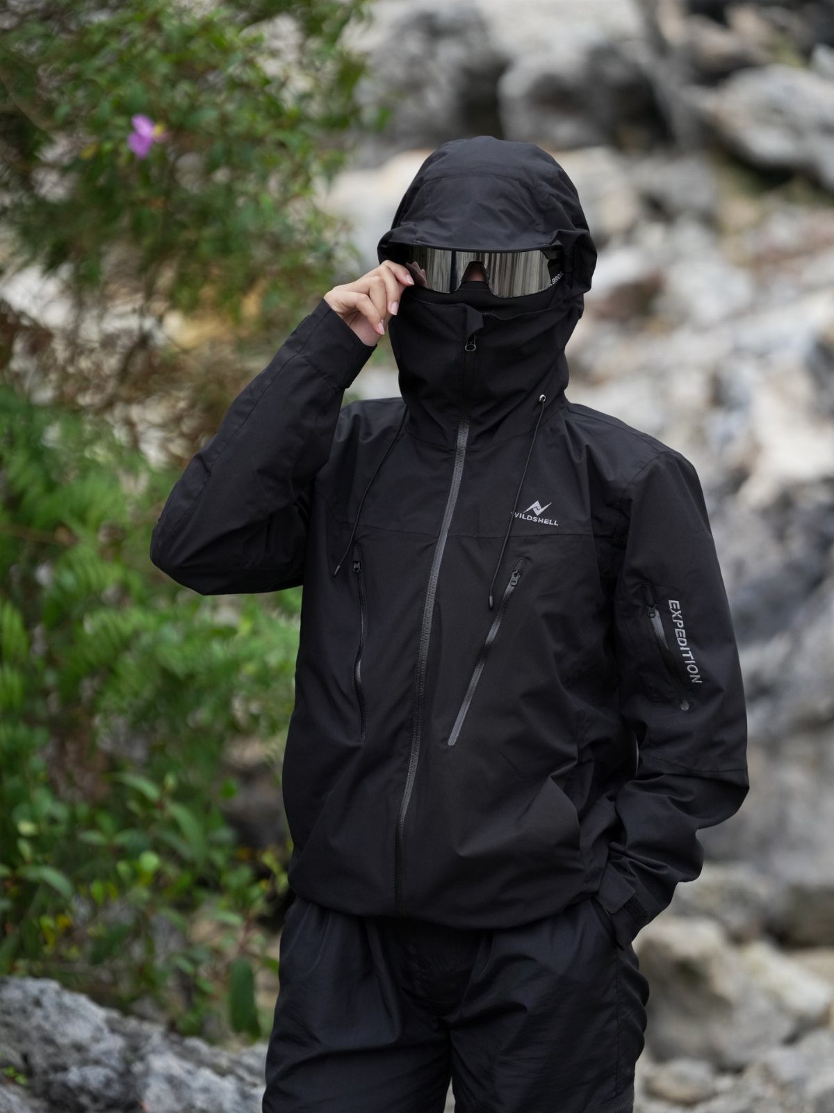 Man in black technical streetwear wearing a hooded jacket and reflective visor goggles outdoors.