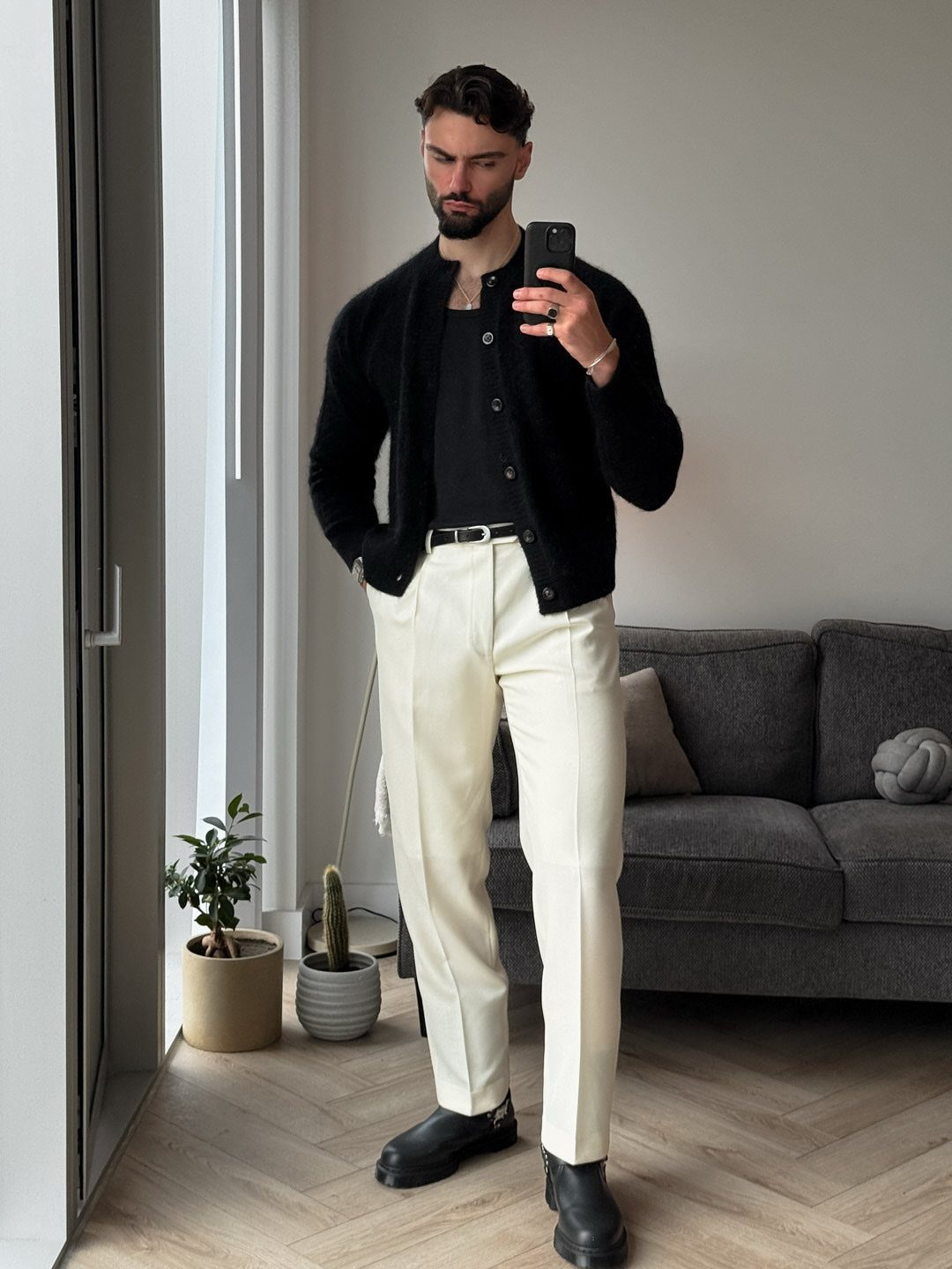 A man posing in a black cardigan, cream trousers, and black boots indoors.