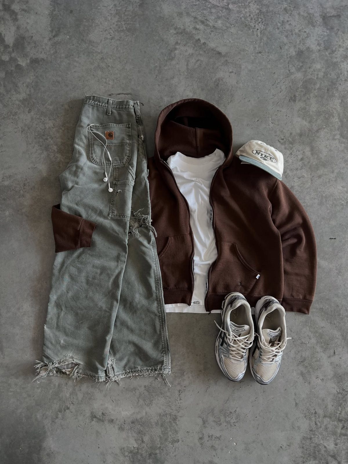 Flat lay of men's streetwear including brown hoodie, white t-shirt, distressed jeans, cap, and sneakers.