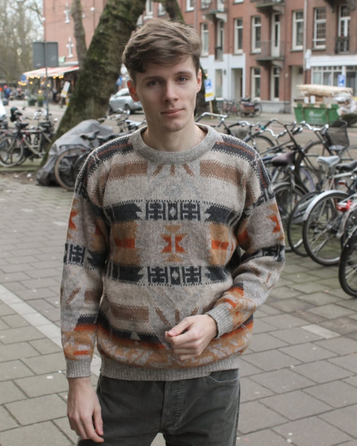 A young man wearing a patterned earth-toned knit sweater standing on a city sidewalk near parked bicycles.