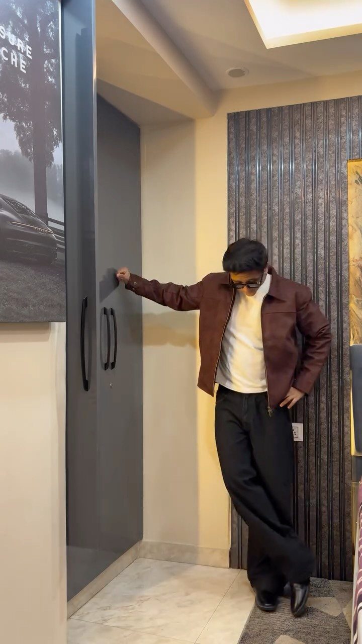 A man wearing a brown structured jacket, white shirt, and black trousers leaning against a cabinet.