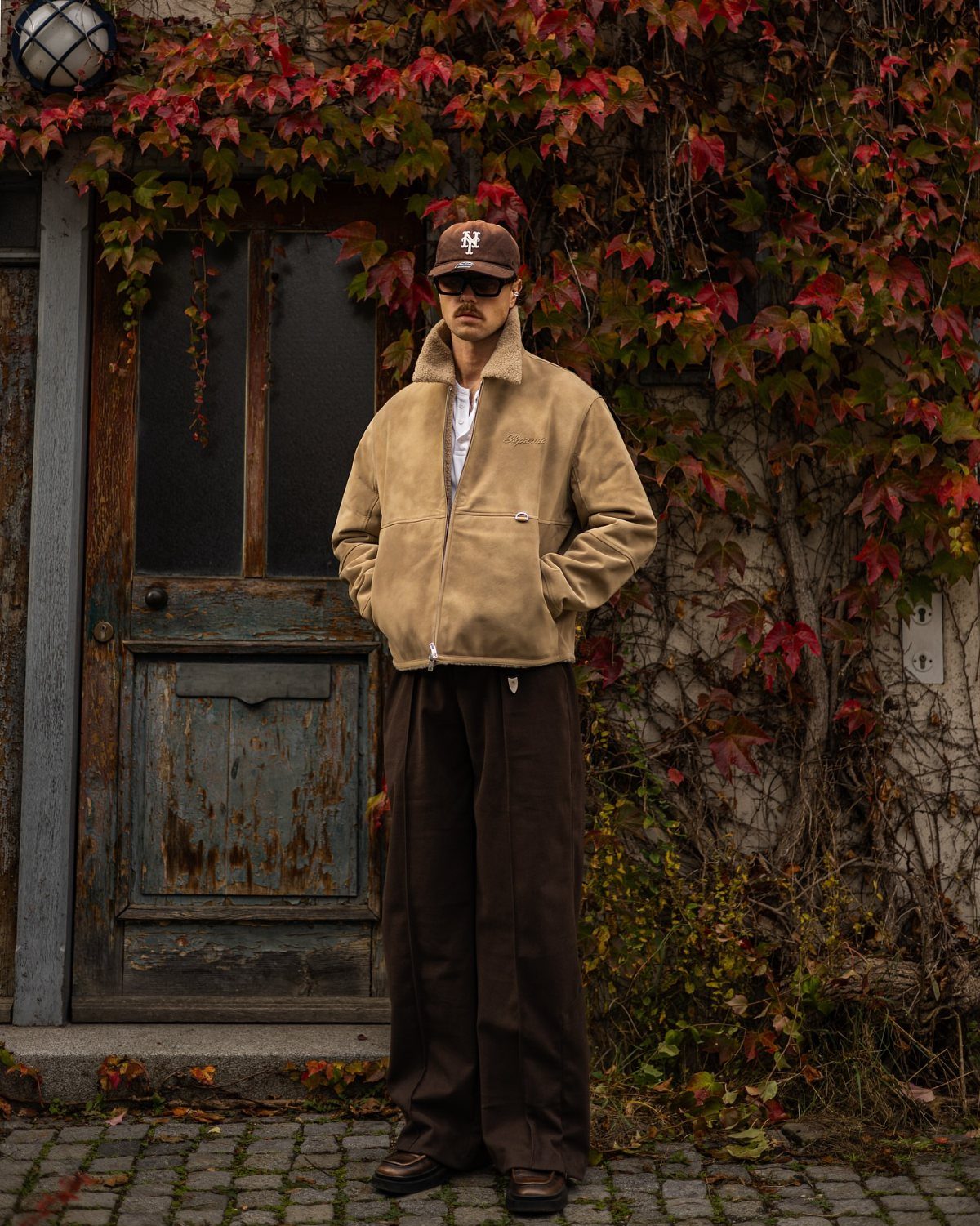 Man wearing a tan jacket and wide-leg trousers standing before a door with red ivy.
