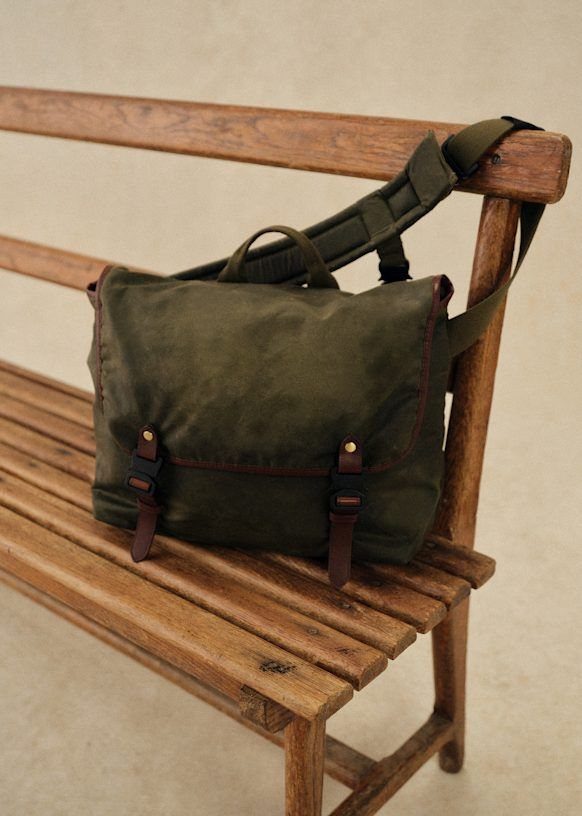 An olive green canvas messenger bag with leather straps resting on a wooden bench.