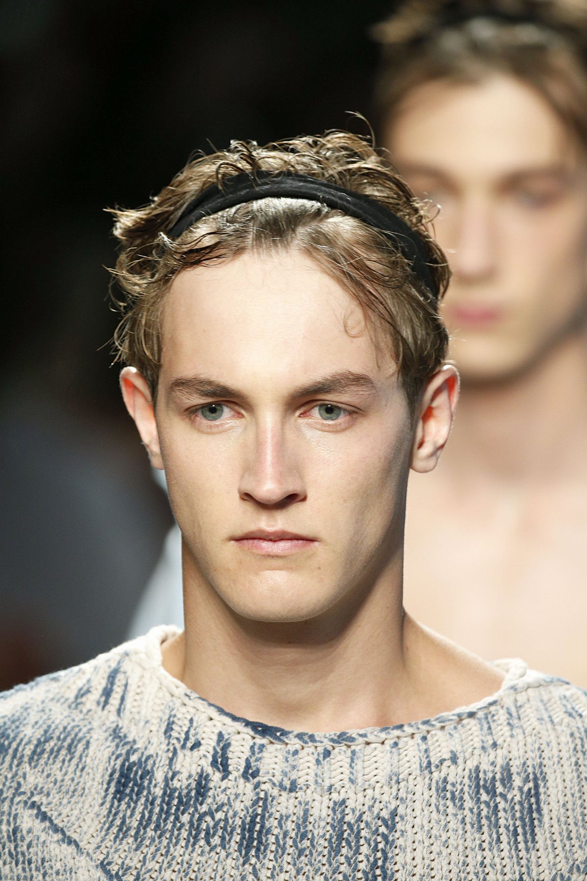 Young man with wavy blonde hair wearing a black headband and a blue and white knit sweater.
