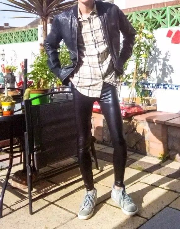 A man wearing a leather jacket, plaid shirt, and shiny black liquid leggings standing outdoors.