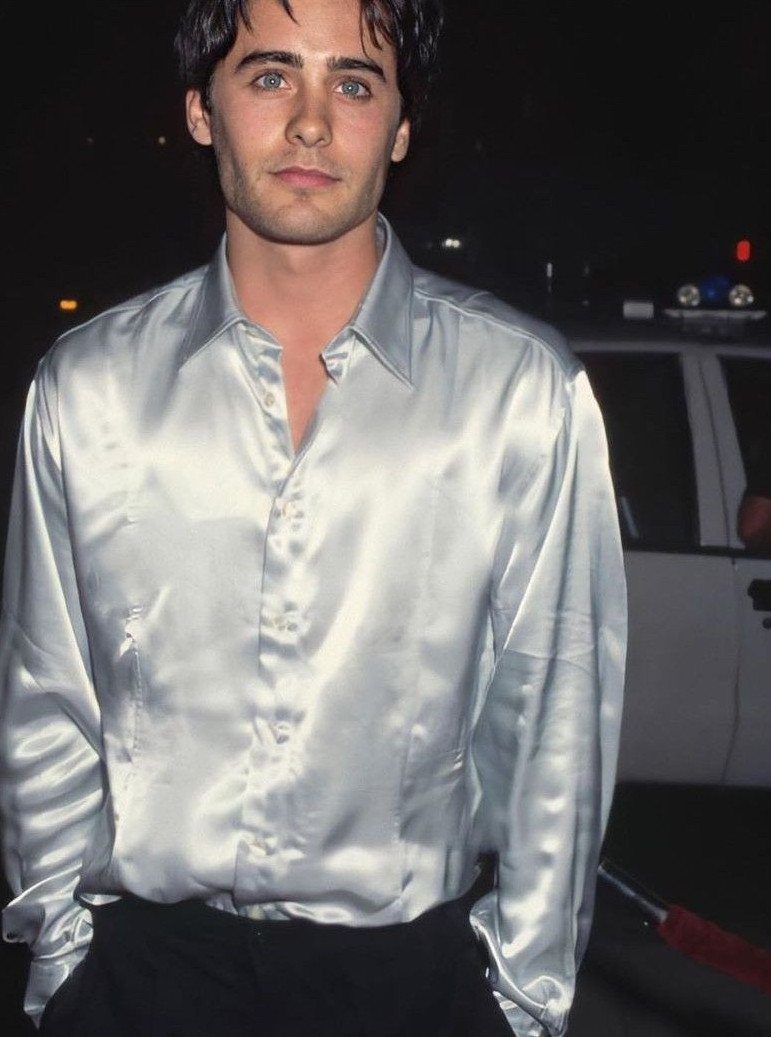 A man wearing a shimmering, light silver satin shirt against a dark night background.